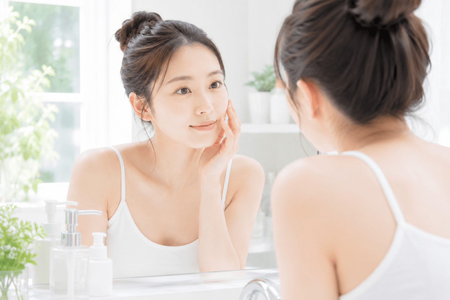 朝の洗面台。窓からの自然光の中で、洗顔前の素肌の女性が鏡をのぞき込み、指先で頬に触れている若い女性