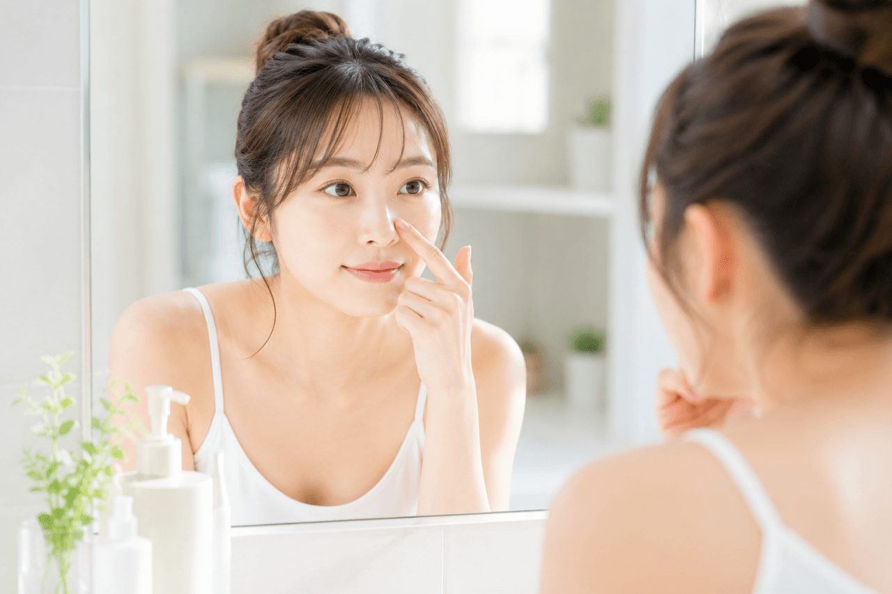 洗面台の鏡の前で、指先で小鼻に触れながら鏡をのぞき込む若い女性。